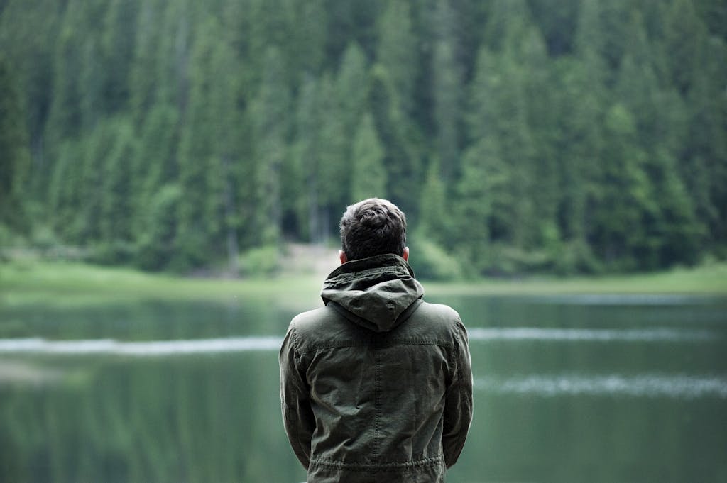 A person in a green jacket stands by a serene forest lake, capturing calming nature.